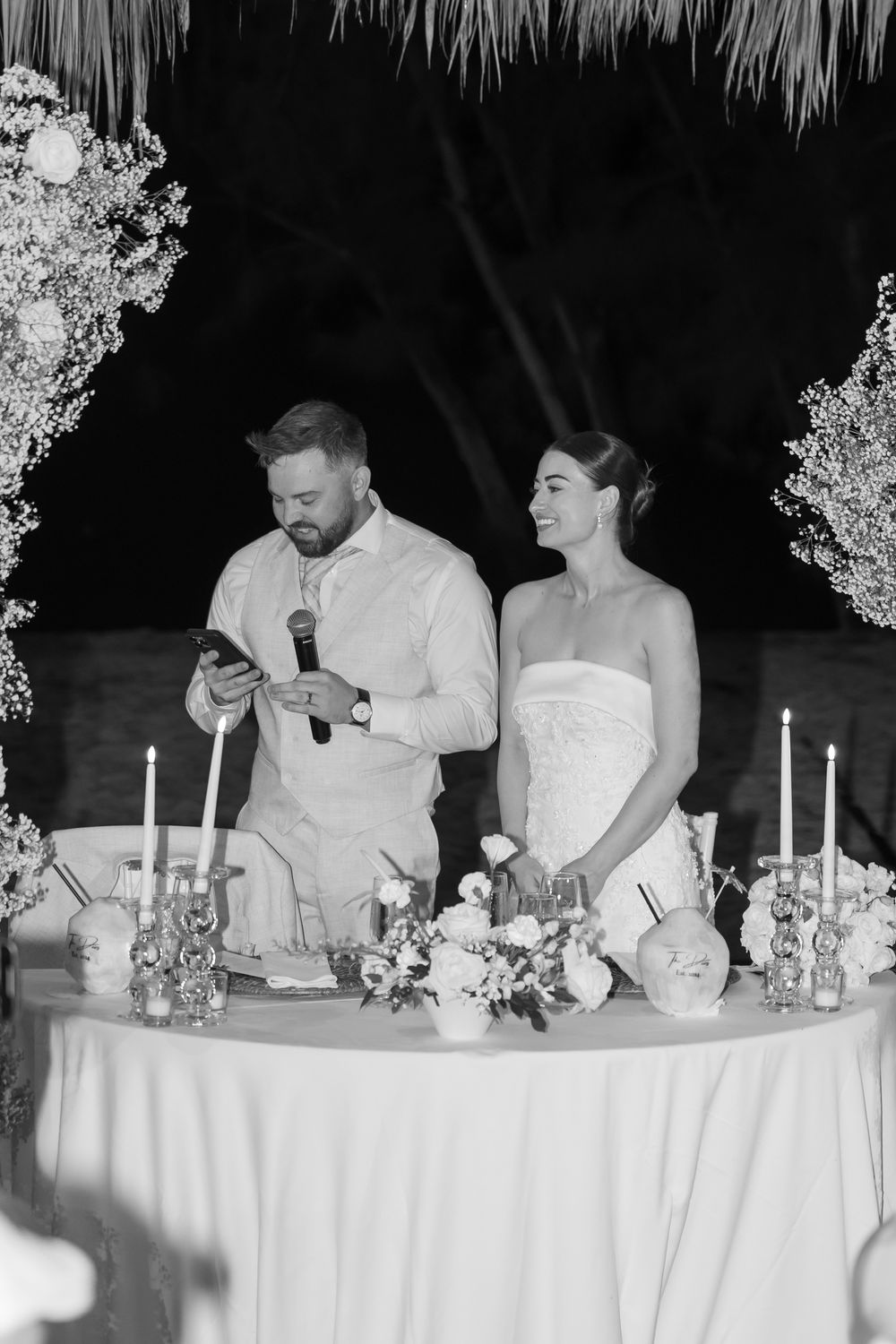 Wedding party seated at an elegant head table with white linens and candlesticks during reception dinner.