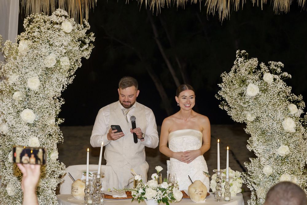 A romantic outdoor wedding reception at night with white floral decorations illuminated by warm lighting.