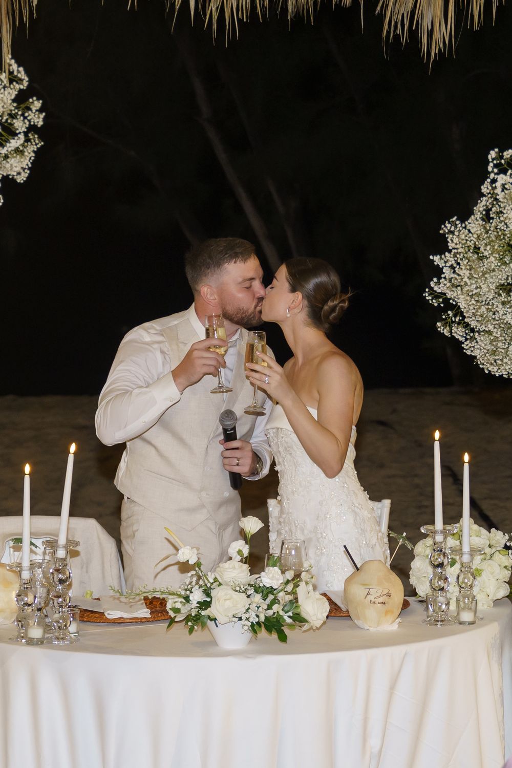 A couple kisses while toasting at their candlelit wedding reception table decorated with white flowers and linens.