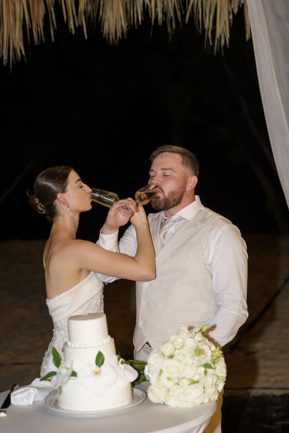 A wedding couple feeds each other cake at their tropical beach reception with hanging decorations.