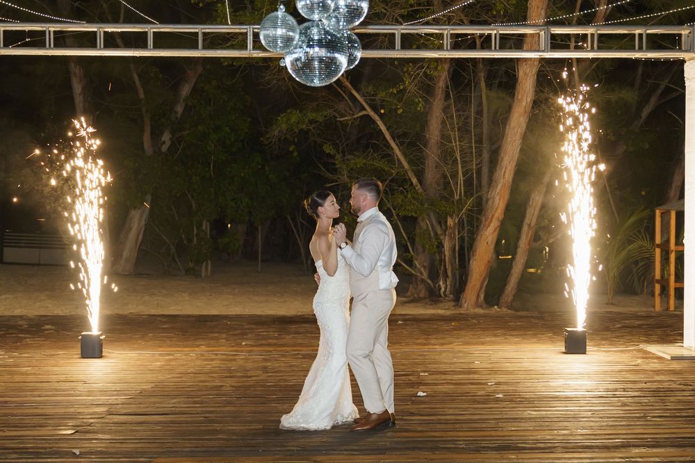 Romantic newlyweds share a tender moment on wooden deck with sparkler fountains and crystal chandelier overhead at night.