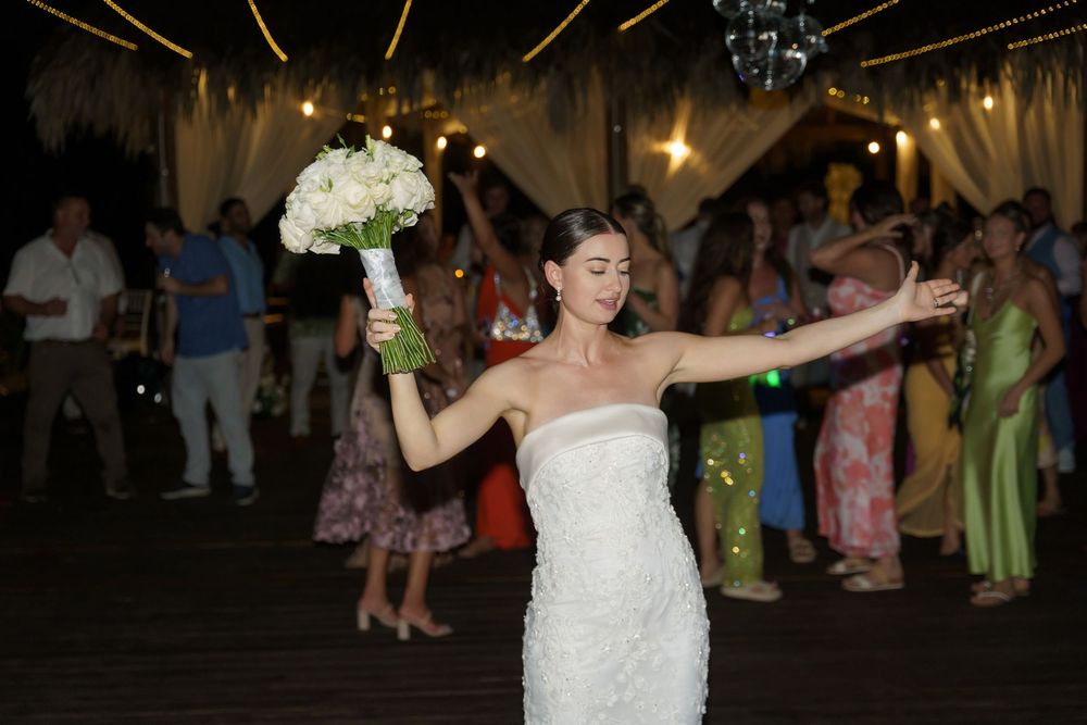 A bride in a white wedding dress tosses her bouquet in the air while other guests watch during the reception.
