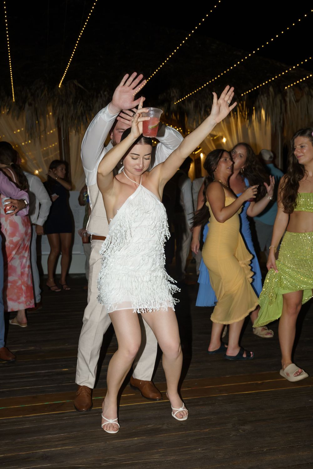 People dance and celebrate on a wooden dance floor under string lights at an evening party.