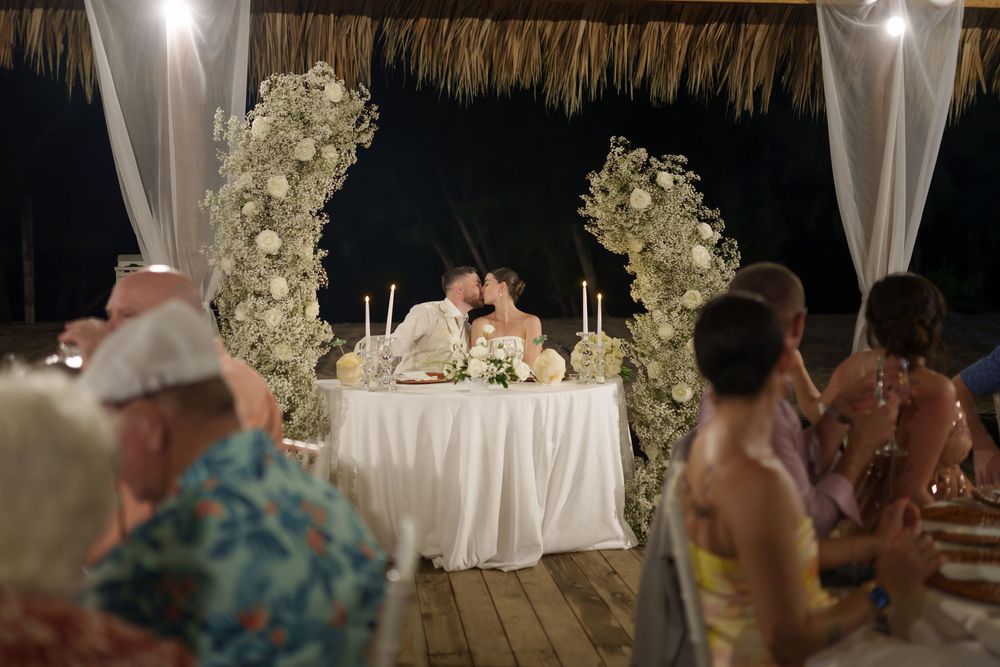 Candlelit wedding reception table decorated with white floral arches on a tropical deck at night.