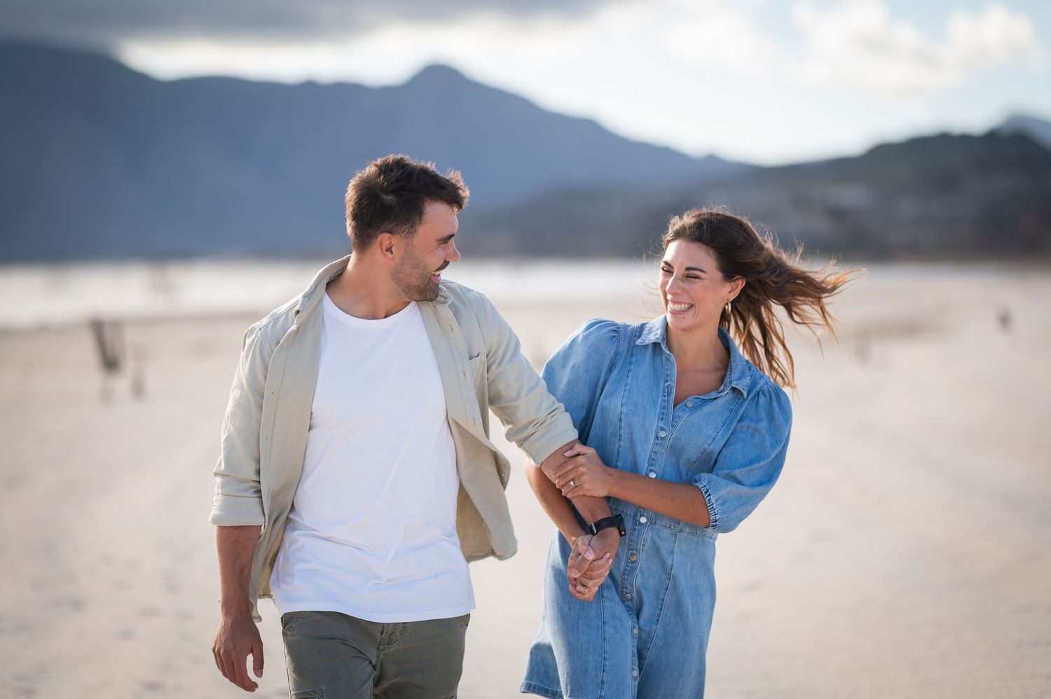 Couple in casual white and denim clothing walking and laughing together on a misty beach.