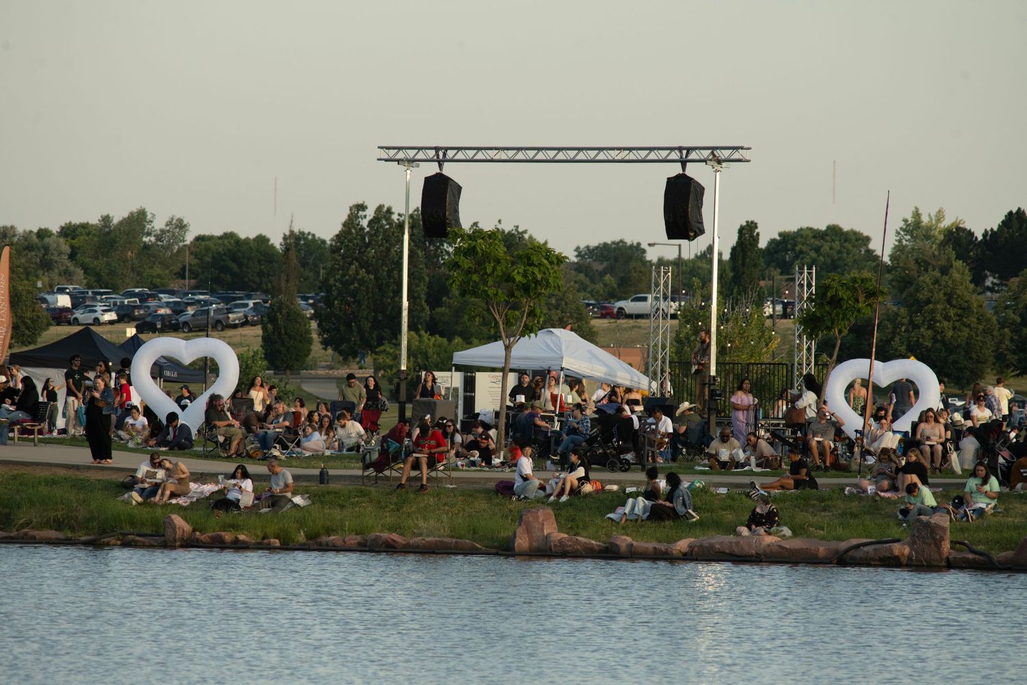 Festival crowd relaxes on grassy waterfront with heart-shaped decorations and speaker systems visible in background.