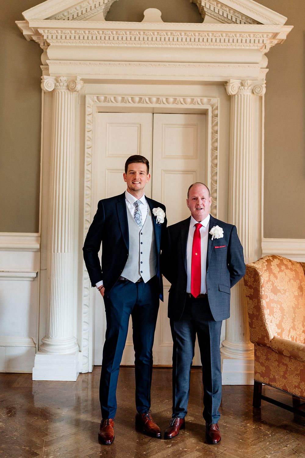 Two people in suits pose in an elegant interior with white columns and architectural details.