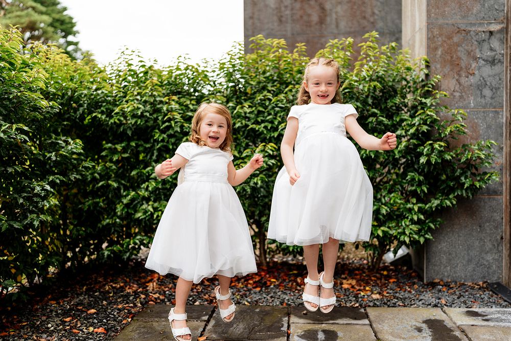 Two excited children in flowing white dresses jumping in a garden setting on a bright summer day.
