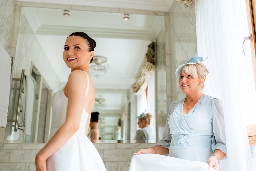 Two people share a joyful moment while getting ready in an elegant hallway with ornate white walls and natural lighting.