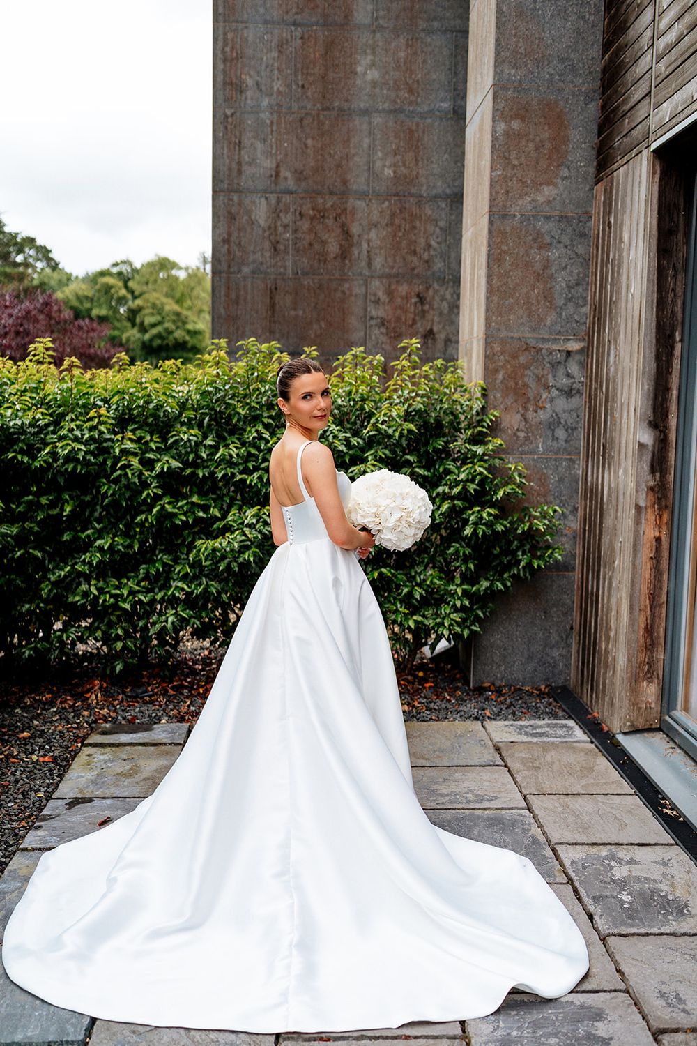 Elegant white wedding dress with dramatic long train photographed against rustic outdoor garden and stone wall setting.