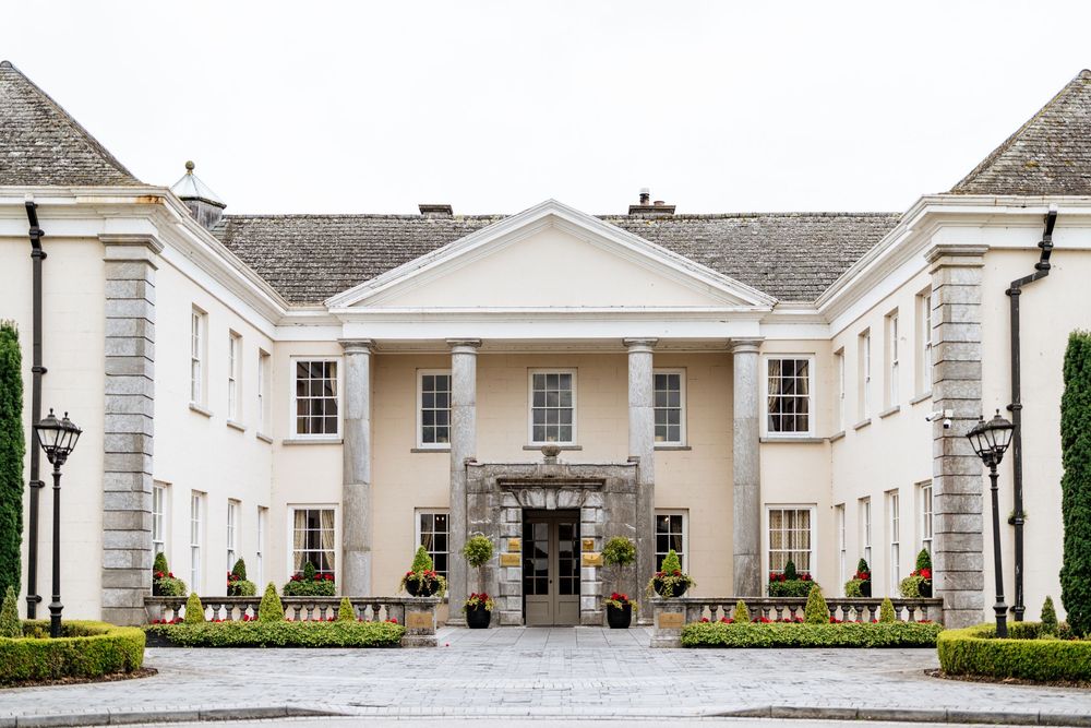 Elegant white manor house with columned entrance porch and symmetrical wings features manicured landscaping.