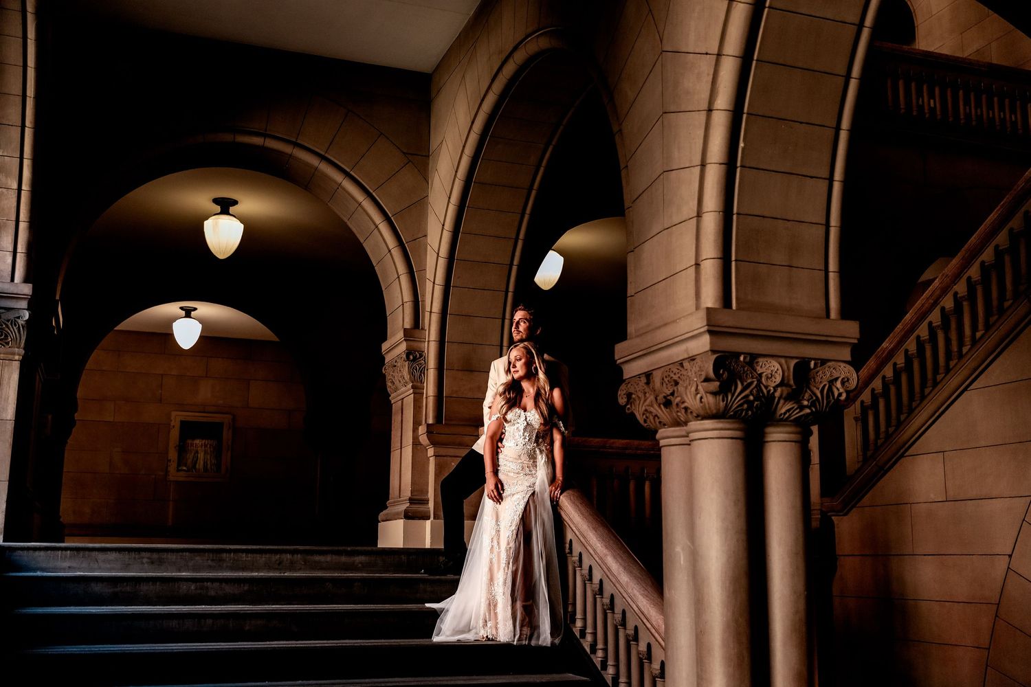An elegant bride in a white gown poses under dramatic arched stone columns with uplighting creating beautiful shadows.