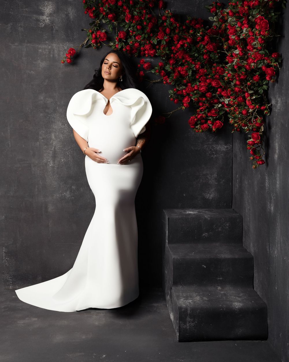 A figure in an elegant white gown with dramatic puff sleeves poses against a dark wall with cascading red flowers.