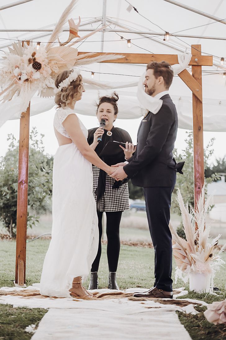 A wedding ceremony takes place under a white tent with dried floral arrangements and wooden beams as decor.