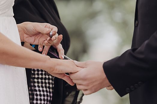 Couple exchanging wedding rings during outdoor ceremony with green trees in the background.