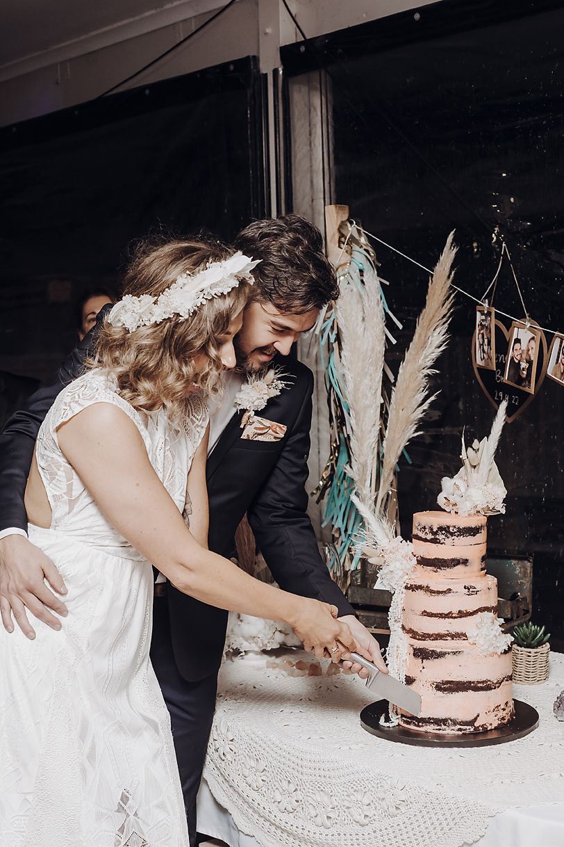 A couple cutting their rustic bohemian wedding cake decorated with dried pampas grass and native florals at reception.