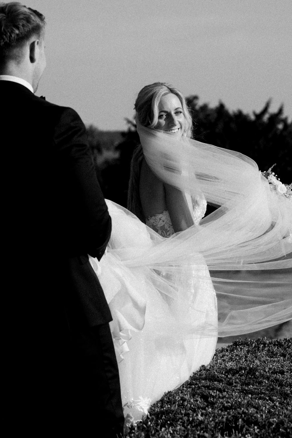 A bride's flowing white wedding veil billows dramatically in the wind against a dark background.