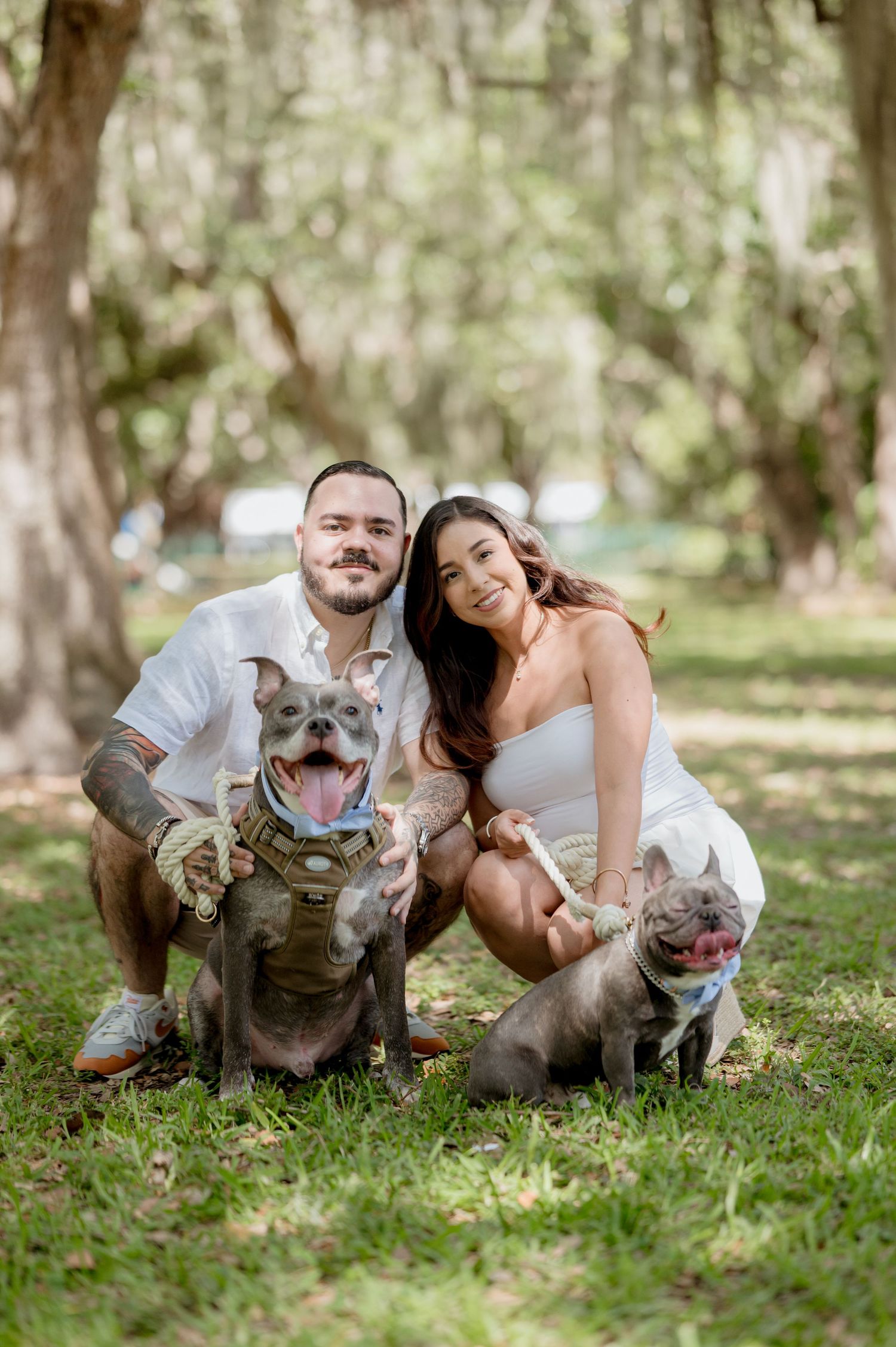 A couple poses outdoors with their two smiling pit bull dogs wearing matching outfits on a sunny day.