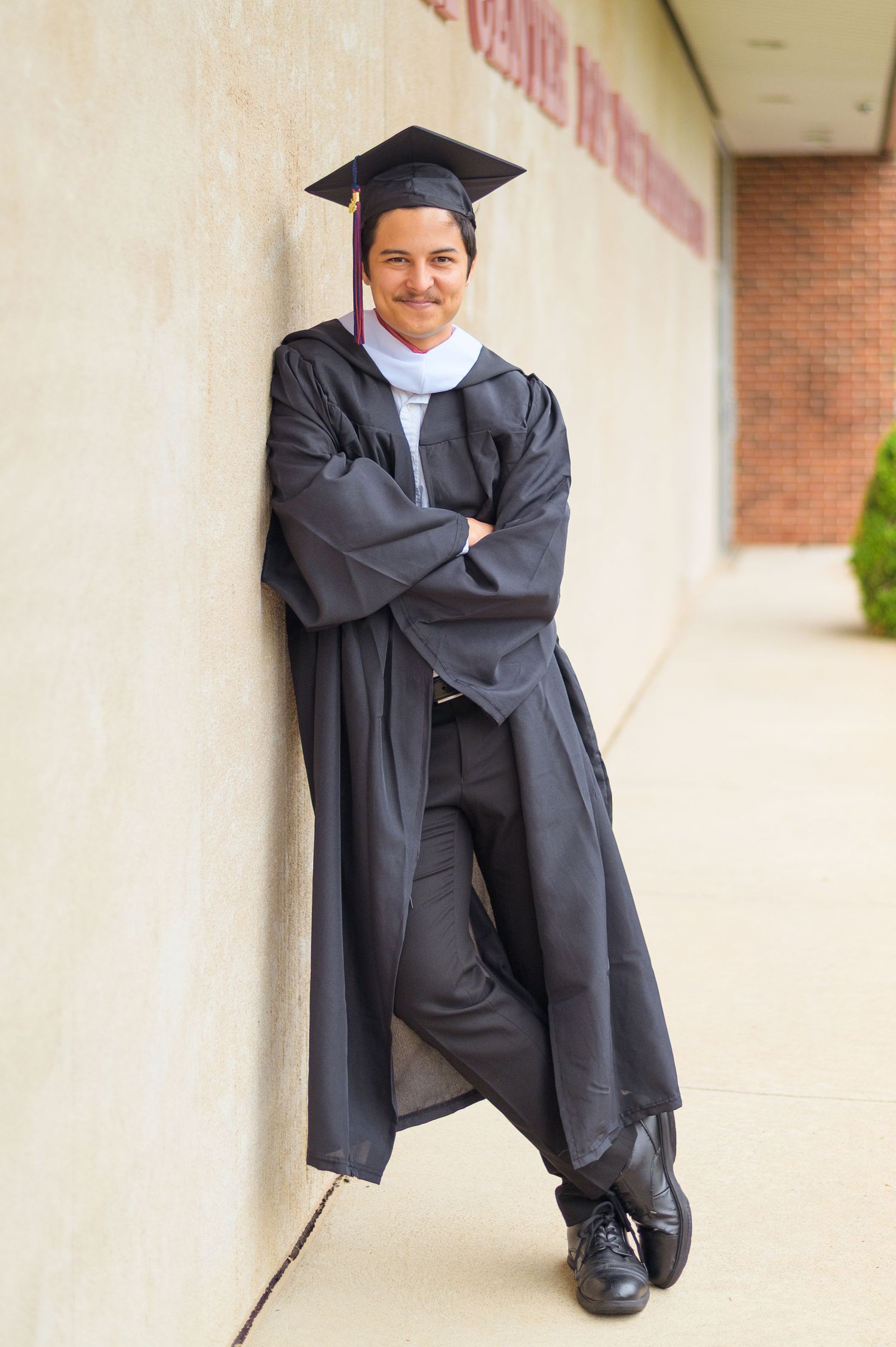 Graduate in cap and gown poses creatively against beige wall.