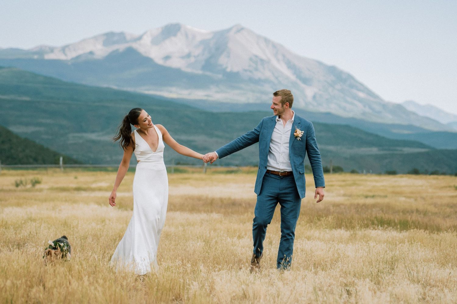 A couple holds hands while walking through a golden mountain meadow with snow-capped Mount Sopris peaks in the distance.