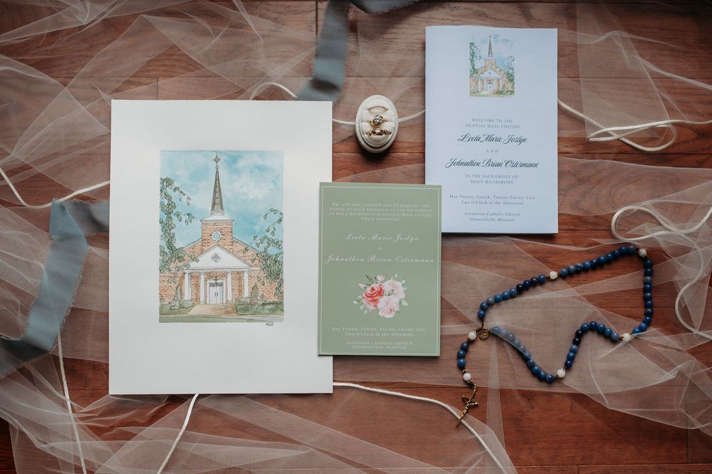 Wedding stationery suite featuring a watercolor church illustration, invitation cards, and rosary beads on a tulle background.