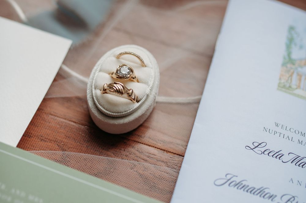 Wedding rings sit in a white oval ring box next to mint green wedding invitation on wooden table.