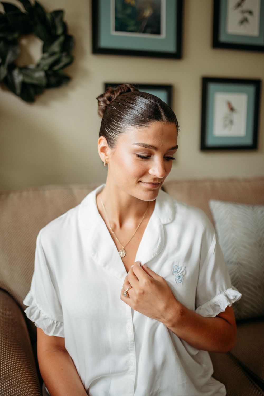 Person in white silk pajama top touching delicate necklace while sitting on brown couch with framed art in background.