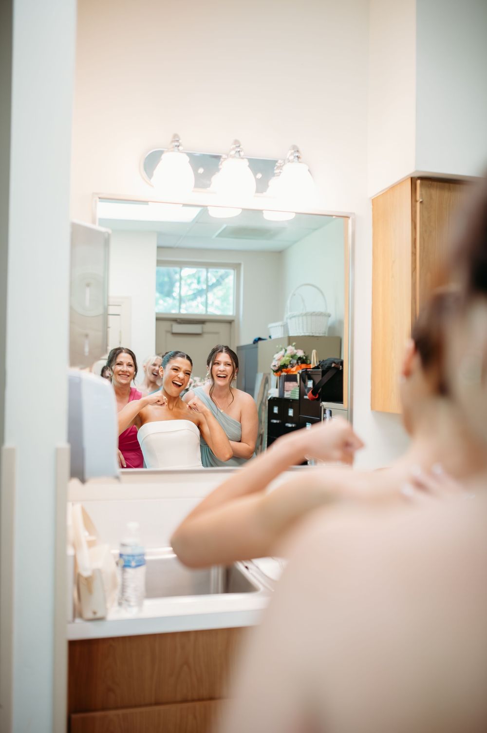 A mirror reflection shows women getting ready in a brightly lit bathroom with white fixtures and wood cabinets.