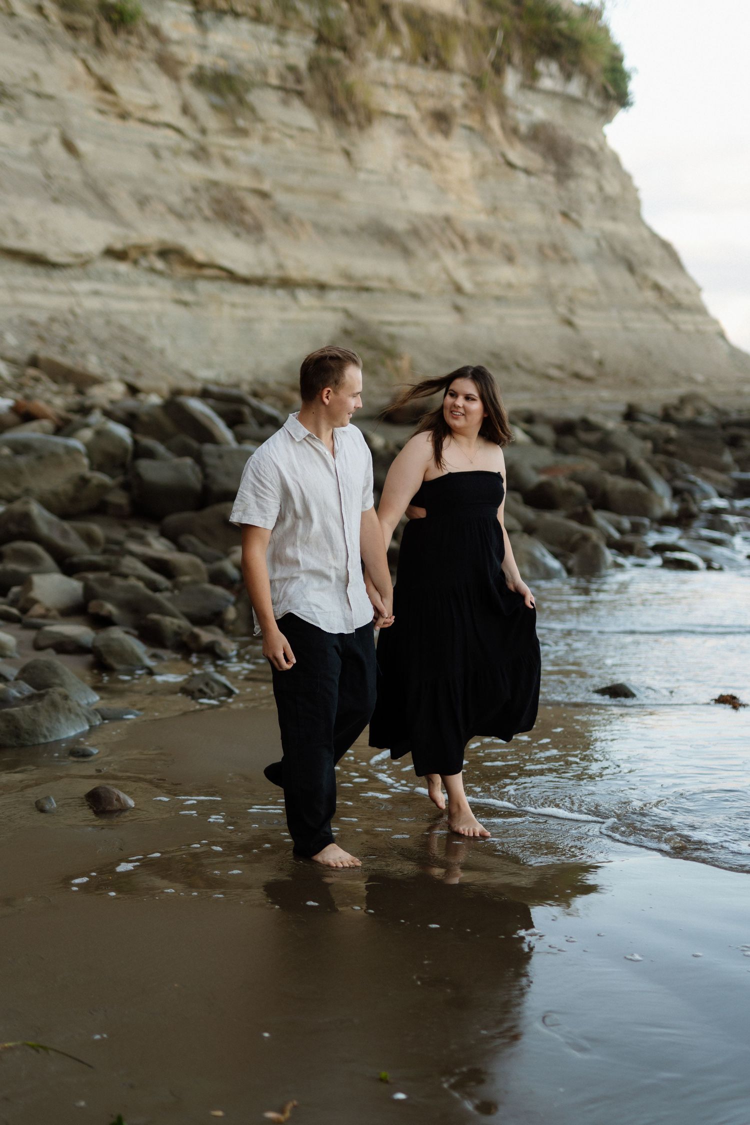 Couple walks barefoot along rocky beach near white cliffside during sunset creating romantic coastal scene.