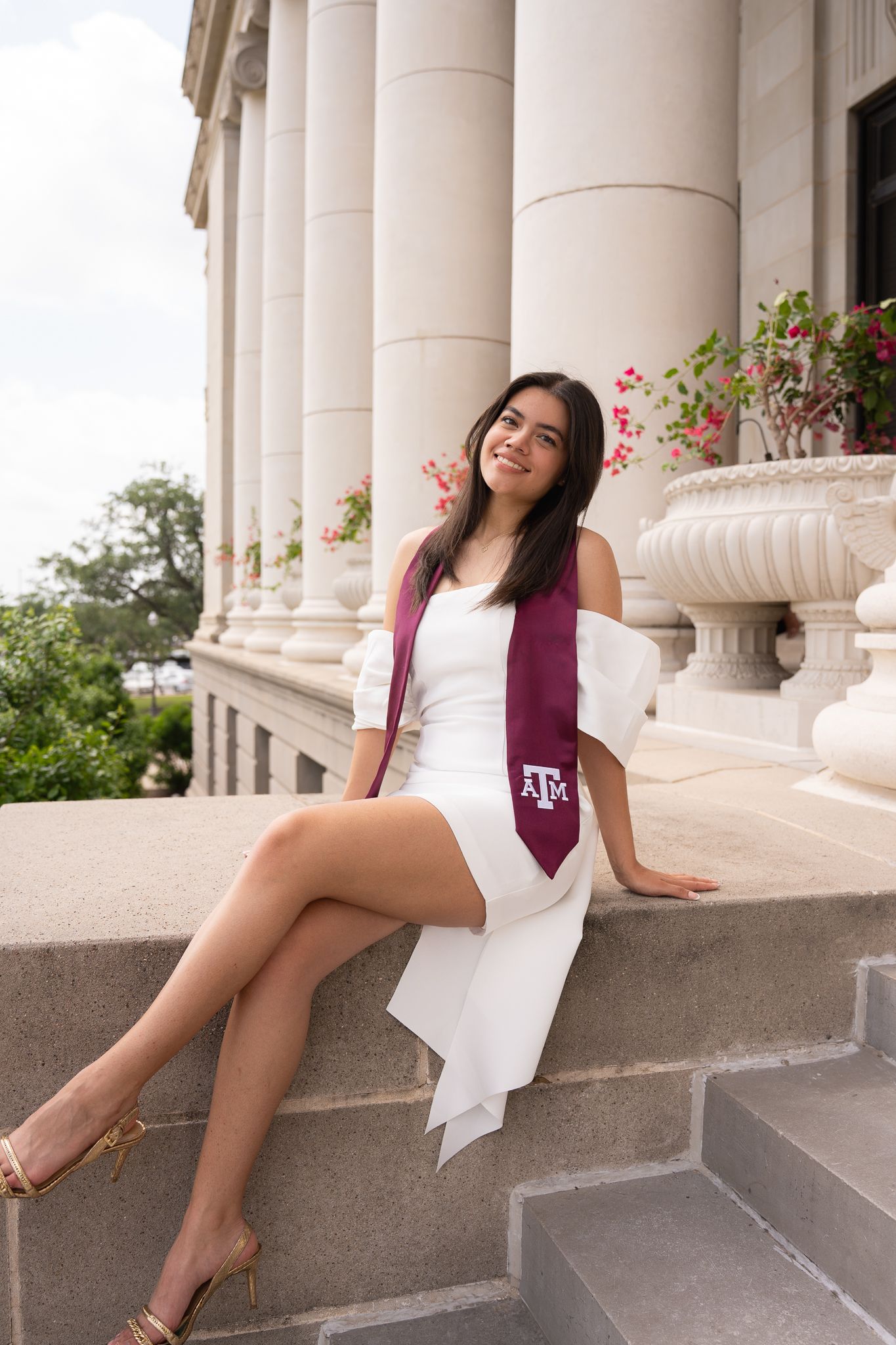 A graduate in a white dress and maroon Texas A&M stole sits on stone steps near classical columns with flowers.