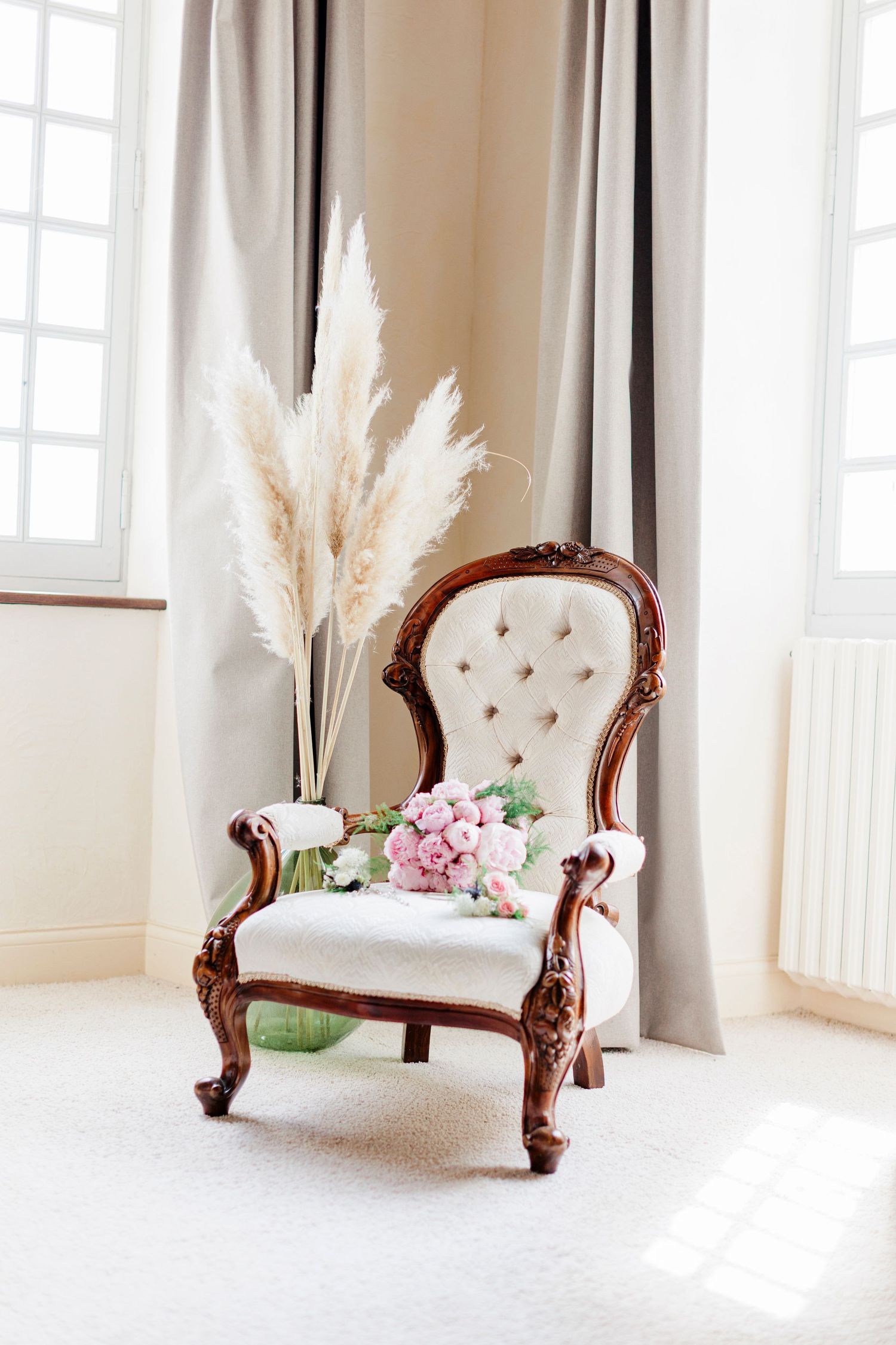 legant vintage armchair with white upholstery and carved wood, decorated with a pink peony bridal bouquet and pampas grass in a bright room