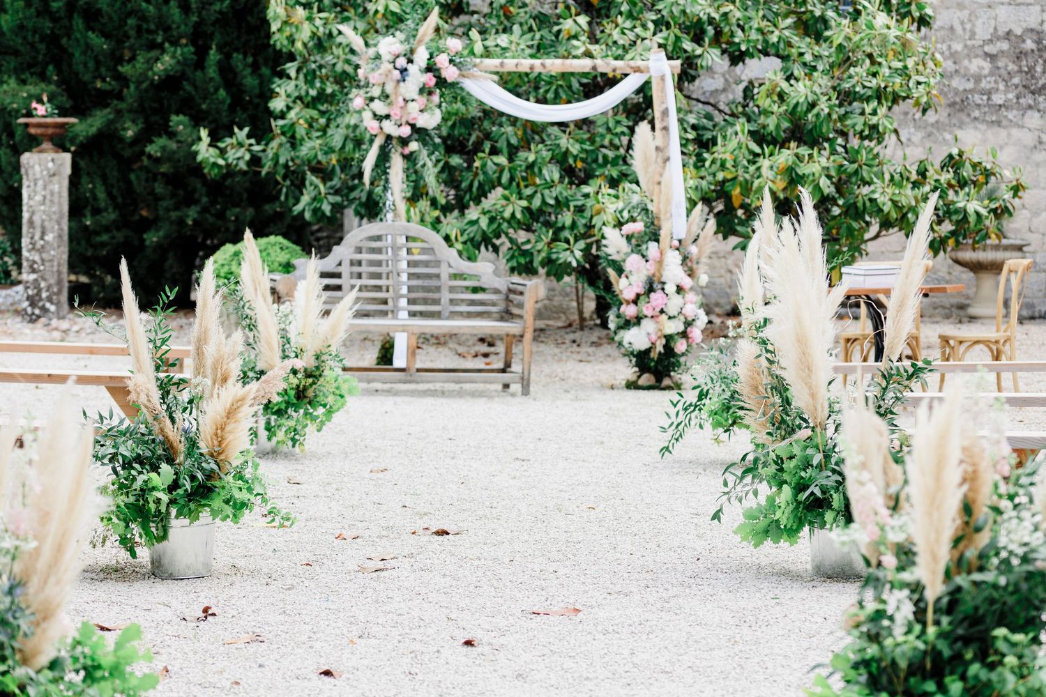 Outdoor wedding ceremony setup at Domaine des Gaillardoux, featuring a floral arch, pampas grass arrangements, and wooden benches under lush