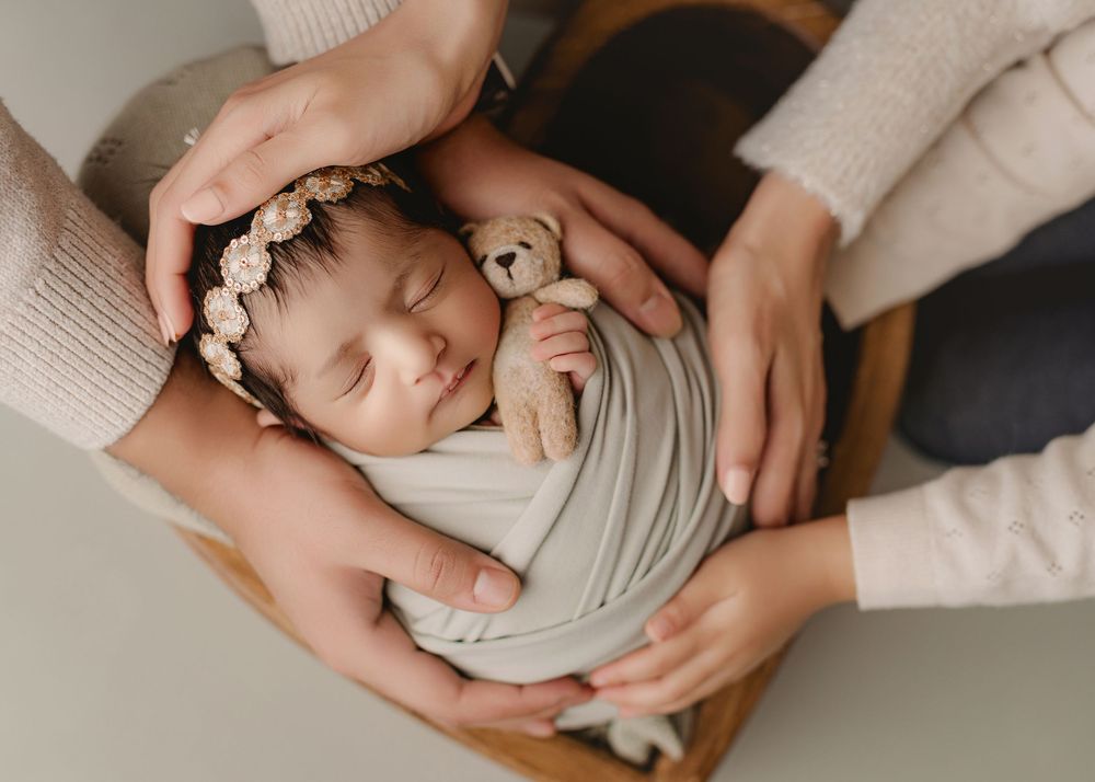 Sleeping newborn wrapped in soft gray fabric swaddle holding small plush teddy bear while cradled in gentle hands.
