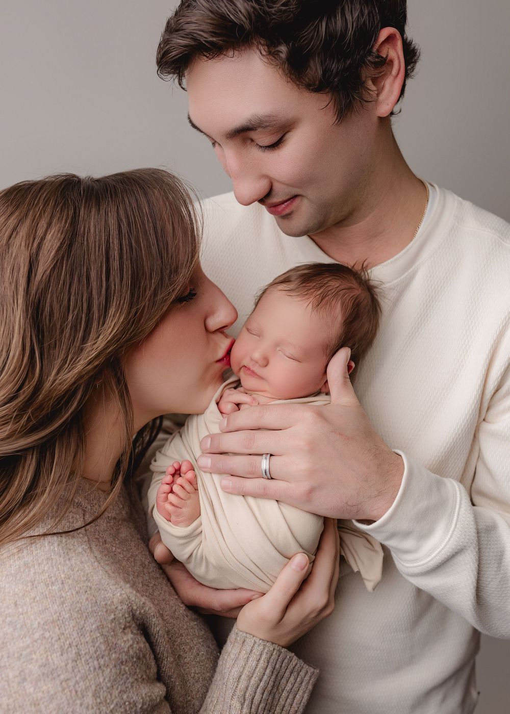 Tender moment between parents and their newborn baby swaddled in white, photographed in gentle neutral tones.