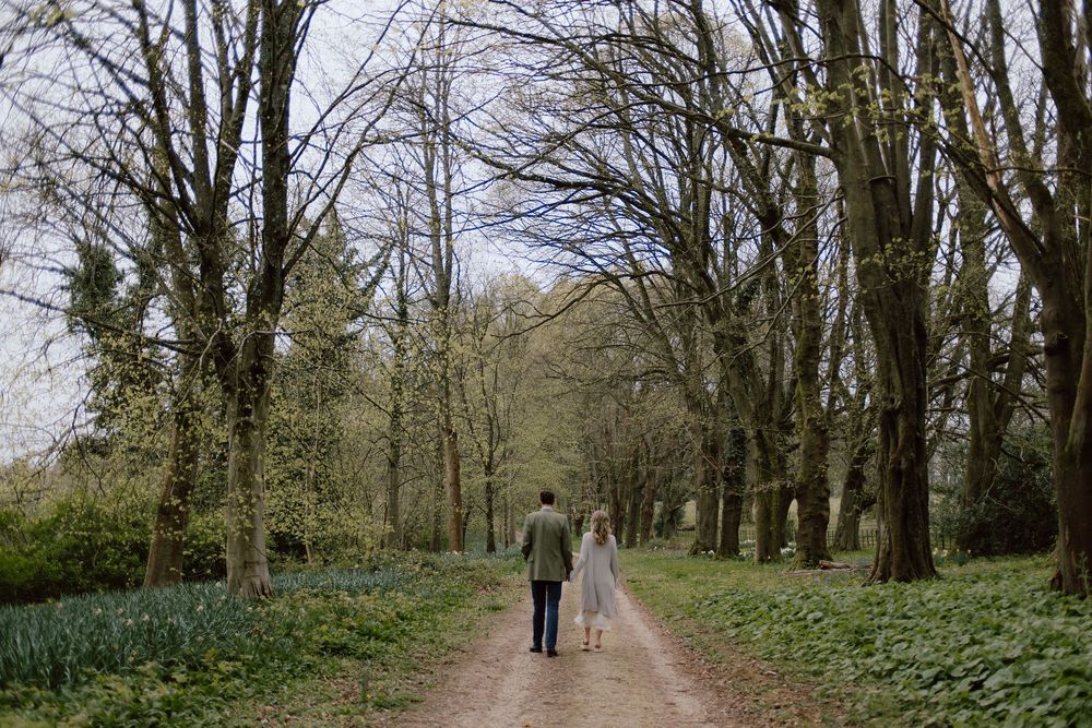 Two people walking with a dog on a scenic tree-lined path surrounded by early spring woodland flowers.