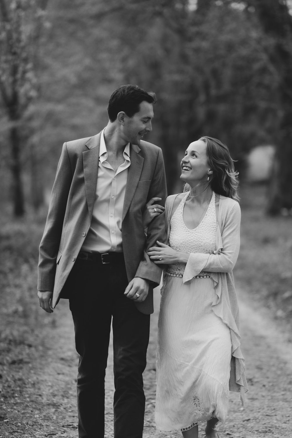 A romantic black and white photo of a couple walking together and sharing a joyful moment on a wooded path.