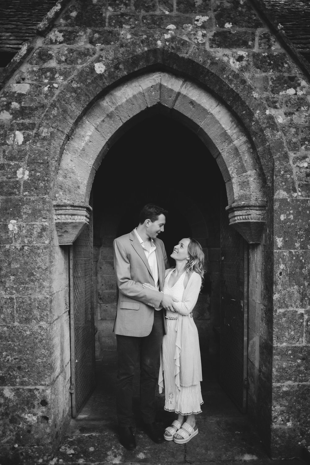 Black and white photo of couple sharing intimate moment under historic stone church archway.