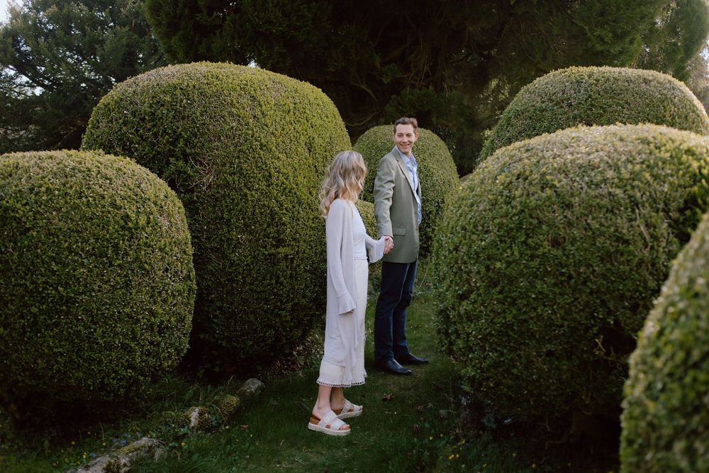 Two figures standing between large rounded topiary bushes in a formal garden at dusk.