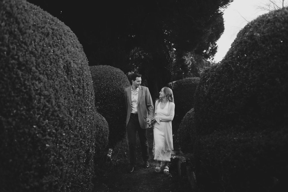A couple stands together in a garden pathway surrounded by tall manicured hedges in black and white.