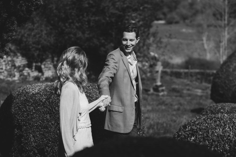 Two people share a tender moment between sculptured hedges in a black and white garden setting.