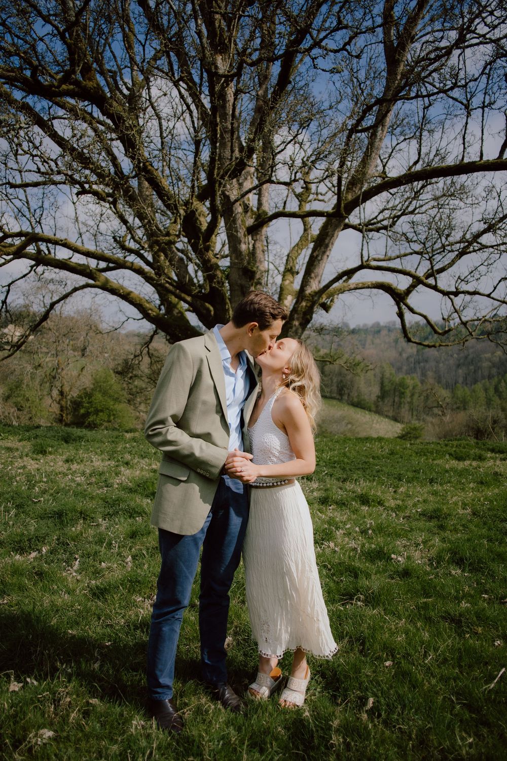 A romantic couple shares an intimate moment under a large bare tree on a grassy hilltop.