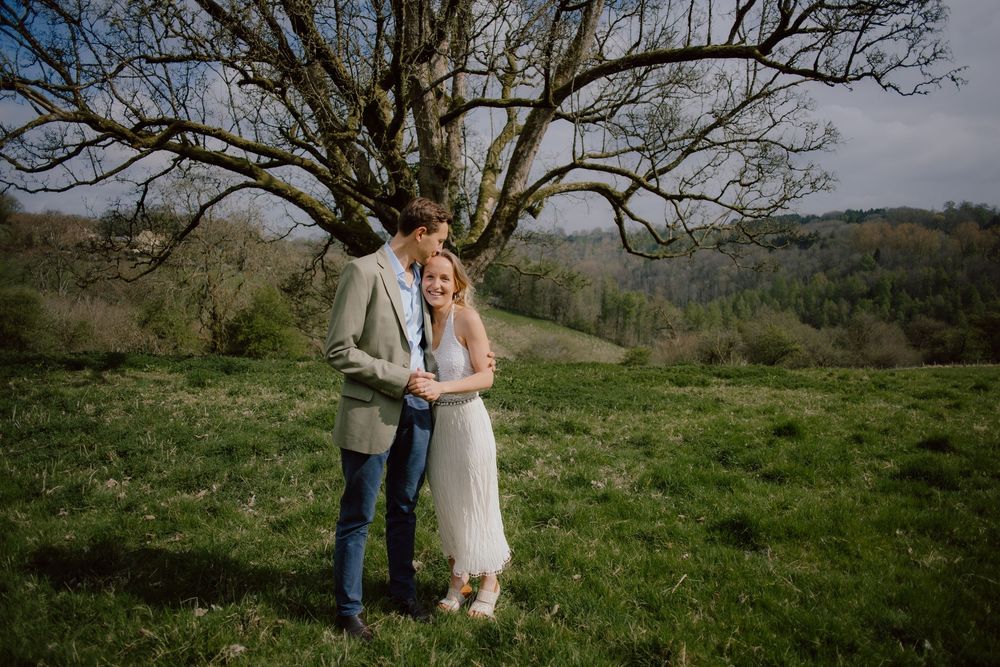 A couple shares a tender embrace on a grassy hillside under a large bare tree in springtime.