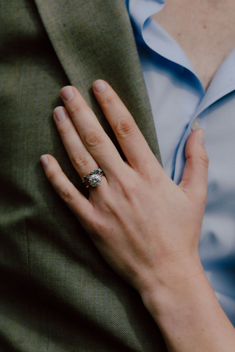 Close-up shot of a hand wearing an ornate engagement ring against olive green and light blue fabric textures.
