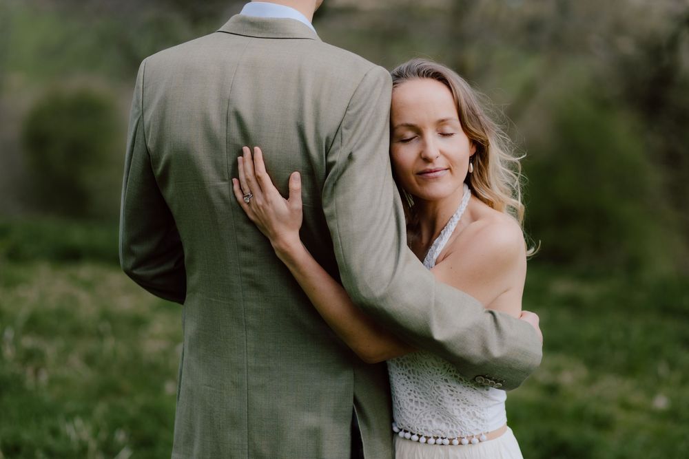 A bride in a white dress embraces her groom from behind in an outdoor setting with greenery.