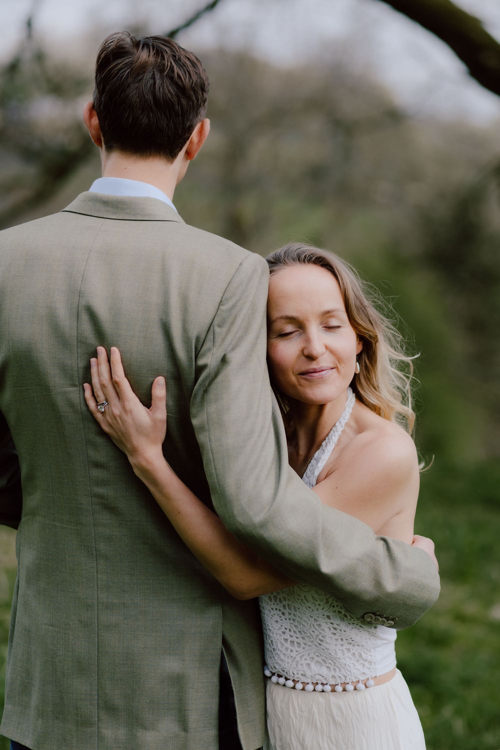 Romantic wedding couple embracing outdoors as bride in lace dress hugs groom in grey suit from behind.