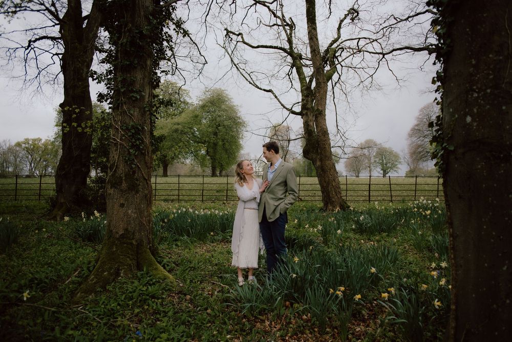 A romantic spring scene in a garden with blooming daffodils and tall trees framing a couple in formal attire.