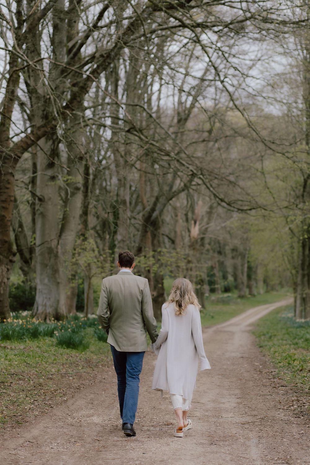 A couple walking hand in hand down a tree-lined dirt path during springtime, wearing light colored clothing.