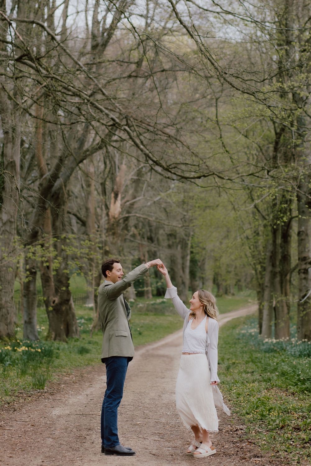 A couple dancing and twirling on a forest path surrounded by budding spring trees.