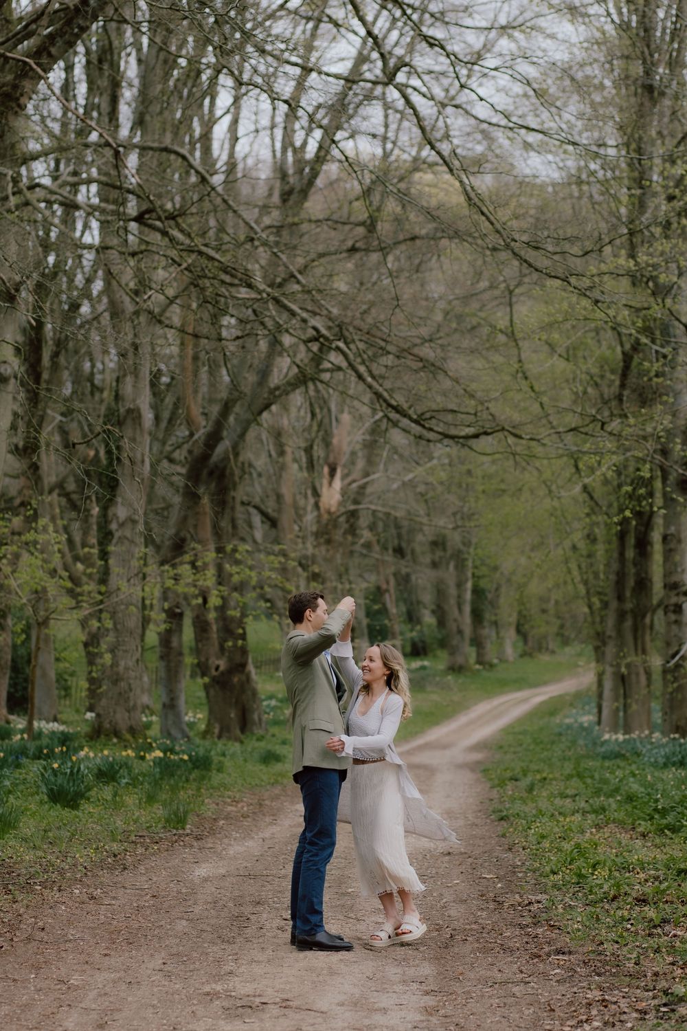 A couple embraces on a tree-lined dirt path with spring flowers blooming in the woodland setting.