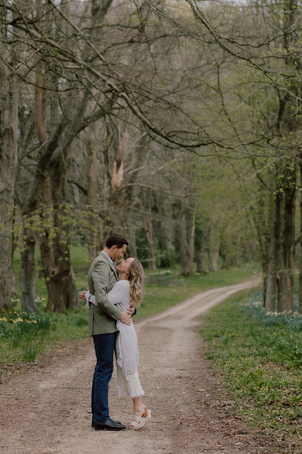 A tender moment between two people embracing on a winding dirt path through a springtime forest with blooming flowers.